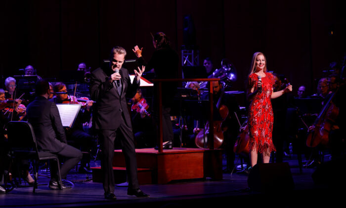 Man in dark suit and woman in red dress singing in front of a symphony orchestra