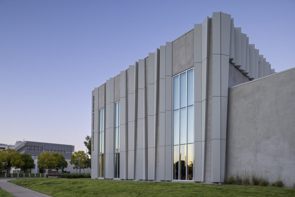 A modern building, the ASO Music Center, with tall vertical windows and textured concrete panels sits on a grassy area under a clear sky. Trees and another building are visible in the background.