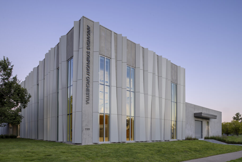 Modern two-story building with vertical white panels and tall windows, labeled ARMENIAN AMERICAN MUSEUM and ASO Music Center, surrounded by grass and trees under a clear sky.