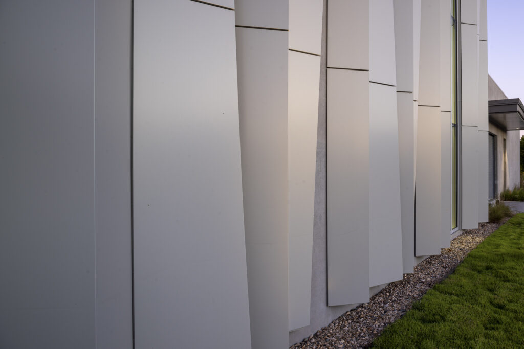 Close-up view of modern building facade at the ASO Music Center, featuring large, angled white panels, a strip of gravel at the base, and a section of green grass on the right.