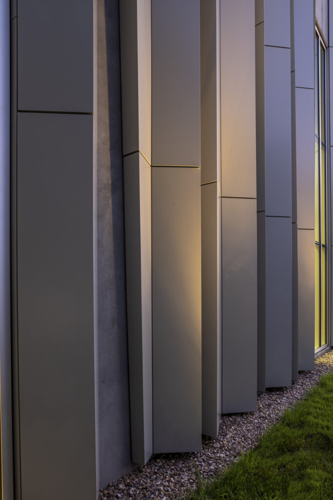 Close-up of the ASO Music Center’s modern facade with vertical gray metal panels, a narrow gravel strip at the base, and a patch of green grass on the right.
