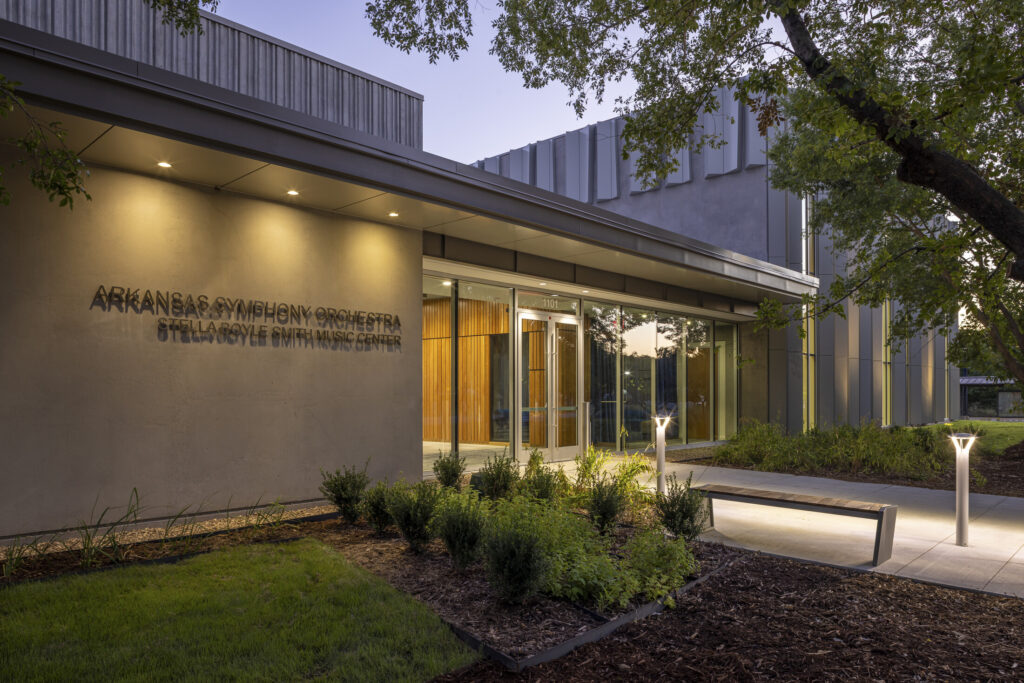 Modern building exterior labeled ASO Music Center and Arkansas Symphony Orchestra Stella Boyle Smith Music Center, with glass doors, foliage, and walkway in early evening light.