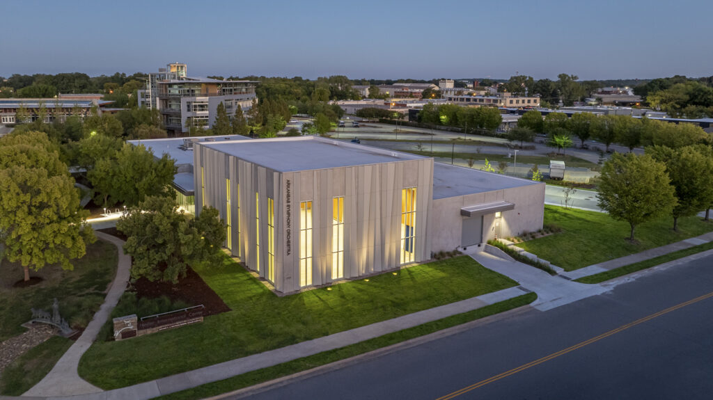 Modern, rectangular ASO Music Center with vertical windows illuminated from inside, surrounded by trees and grass, located at a street corner in an urban area at dusk.