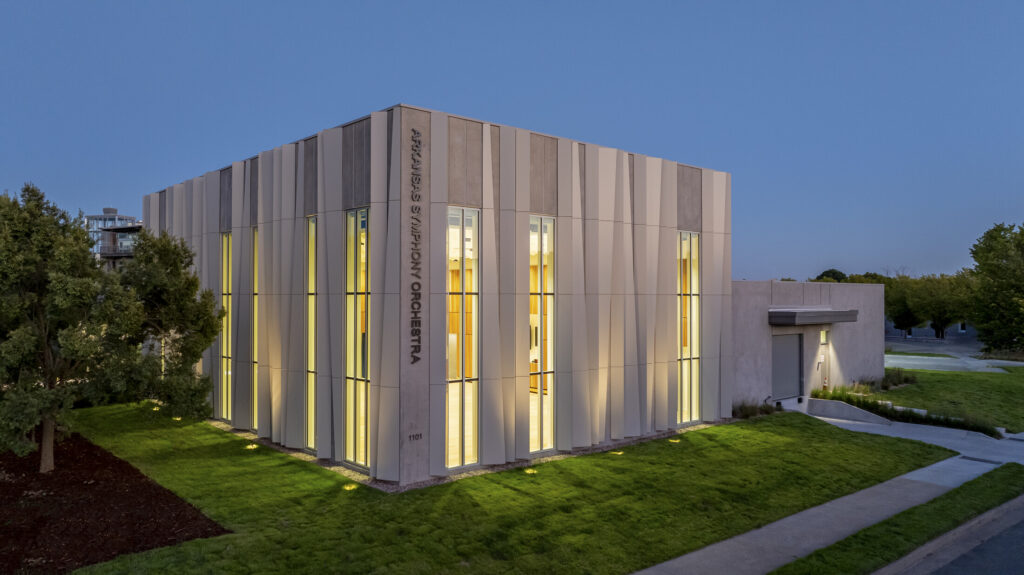A modern building with tall vertical windows, illuminated from within at dusk, labeled ASO Music Center on the exterior wall, surrounded by grass and trees.