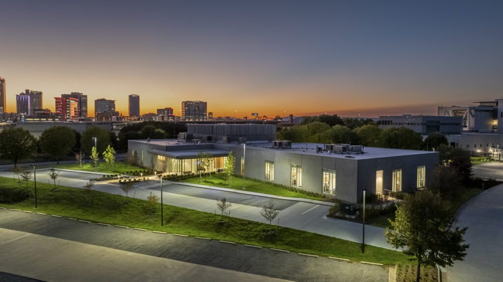 A modern, single-story building with large windows glows at dusk—the ASO Music Center—framed by trees and a parking area, with a city skyline visible in the background.