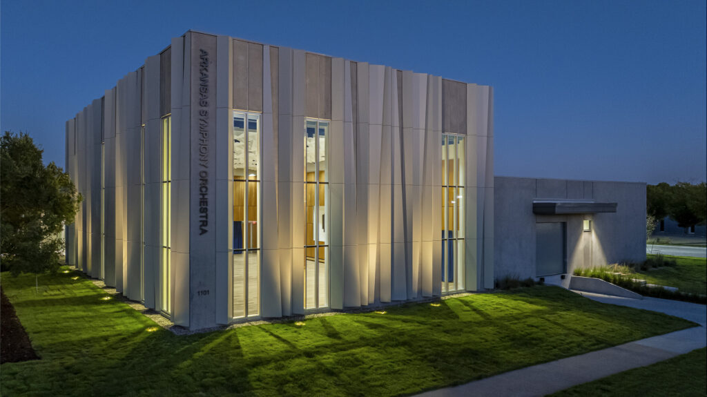 A modern building with ASO Music Center and Arkansas Symphony Orchestra written vertically on the facade, illuminated at dusk with large vertical windows and a landscaped green lawn.