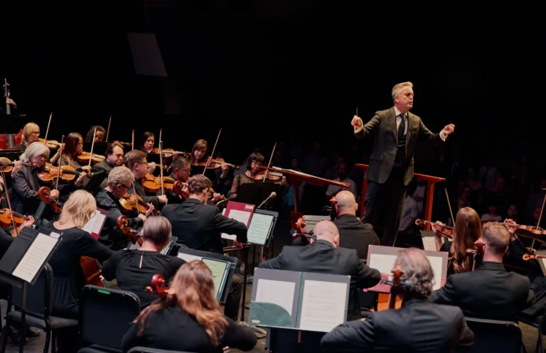 A conductor leads the ASO orchestra of musicians playing string instruments during a live River Rhapsodies performance on stage.