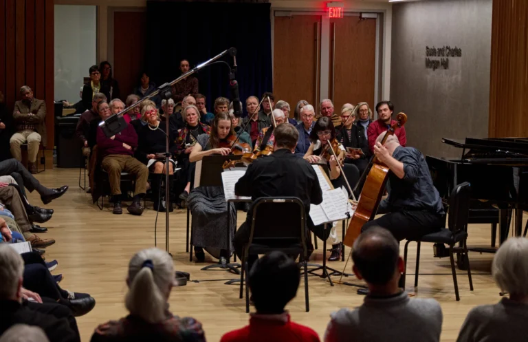 A string quartet performs on stage for an attentive seated audience in a recital hall, with a microphone overhead and a piano visible on the right at this captivating chamber concert.