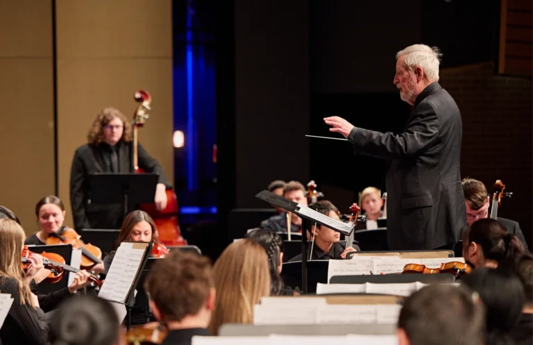 Tom McDonald leads the Academy Orchestra during a performance, with musicians playing string instruments and reading sheet music.